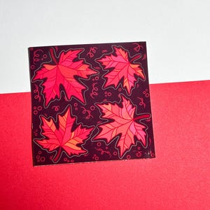 May include: Square sticker with four stylised red and orange maple leaves on a dark background. The leaves have detailed veins and are outlined in white. The background has small, decorative swirls.