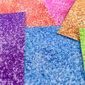 Può includere: Primo piano di fogli di carta glitterata colorata nelle tonalità di viola, rosso, rosa, oro, blu e verde. La carta ha una superficie strutturata e scintillante, ideale per lavori creativi.