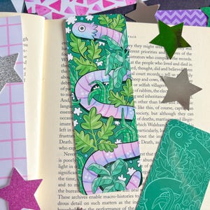 May include: A rectangular bookmark featuring a light purple snake with dark purple stripes winding through a dense pattern of green leaves and foliage. The bookmark is placed on an open book page surrounded by various colorful star-shaped cutouts.