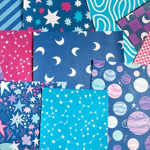 May include: A selection of patterned papers with celestial themes. The papers showcase diverse designs, such as constellations, stars, moons, planets, and wavy lines. The colour palette includes blues, pinks, purples, and white. The papers are arranged in a layered, overlapping composition.