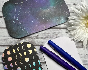 May include: Three blank note cards with rounded corners. One card has a black background with a holographic moon phase pattern. Another card has a dark blue background with a constellation pattern. The third card is white.