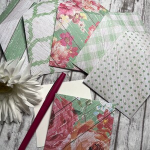 May include: A set of five decorative envelopes with floral and geometric patterns on a whitewashed wood background. The envelopes are green, white, and pink with a distressed look. A red pen and a blank card are also included.
