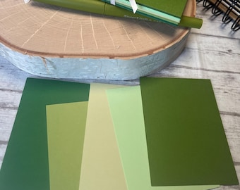 May include: A stack of green cardstock paper in various shades of green, including dark green, light green, and a pale yellow-green. The paper is tied together with a white ribbon and is sitting on a wooden surface.