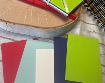 May include: Five blank note cards in different colors: red, light blue, white, navy blue, and lime green. The cards are arranged in a row on a wooden surface.