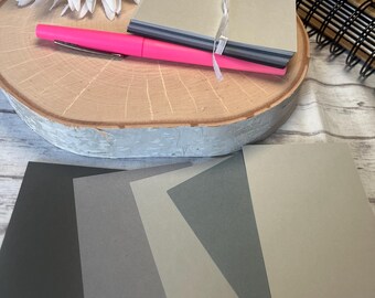 May include: Four sheets of cardstock in different shades of gray, including black, dark gray, light gray, and a very light gray. The sheets are arranged in a row on a wooden surface.