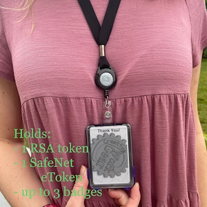 May include: A black lanyard with a retractable badge holder. The badge holder has a clear plastic front and a gray back with the text "Thank You! Creative Gear Work" and a gear design. The text "Holds: 1 RSA token, 1 SafeNet eToken, up to 3 badges" is written in green text below the badge holder.