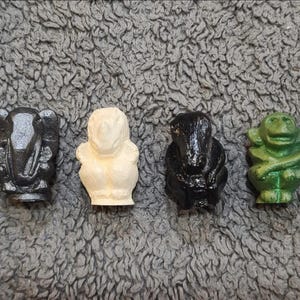 May include: Four small, molded figurines arranged in a row. The first is a black elephant, the second is a cream-colored rhinoceros, the third is a black creature with a long snout, and the fourth is a green monkey with its arms crossed.