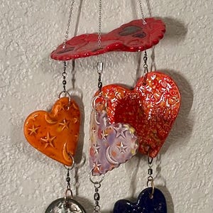 May include: A hanging mobile featuring heart-shaped ornaments in various colors, including red, orange, purple, and blue. The hearts have textured surfaces and star patterns, suspended by chains.