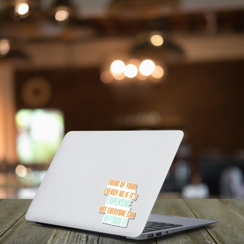 May include: A white laptop computer with a sticker that reads "THINK OF YOUR ENERGY AS IF IT'S EXPENSIVE, NOT EVERYONE CAN AFFORD IT" in orange, teal, and white text. The laptop is on a wooden table with a blurred background.