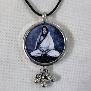 Puede incluir: Un collar con un colgante redondo de plata con una imagen de una mujer con un sari blanco. El colgante tiene un borde de plata y una pequeña cadena de plata con tres cascabeles de plata colgando de él.