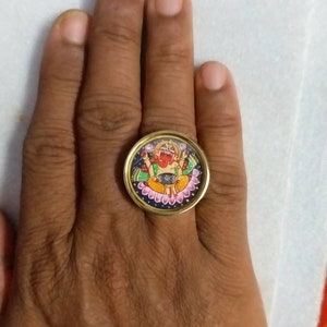 May include: A gold ring with a round, colorful, painted design of a deity with multiple arms. The deity is sitting on a lotus flower and has a red face.