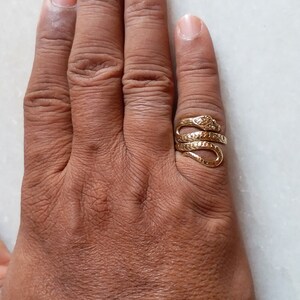 May include: A gold-toned snake ring with a textured surface. The ring is worn on the middle finger of a hand.