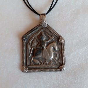 May include: Silver pendant necklace with a detailed image of a person riding a horse. The pendant is in a rectangular shape with a decorative border.