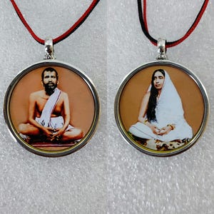 May include: Two round pendants with images of people, each in a silver-colored setting. The pendants hang from a black and red cord. The images are of people in traditional clothing, seated in a meditative pose.