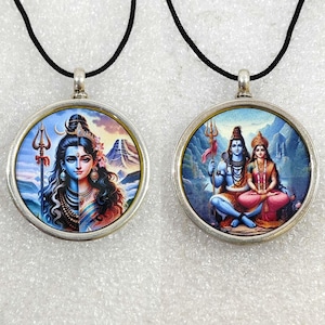 Puede incluir: Dos colgantes de plata con cordones negros. Un colgante presenta una imagen colorida de Shiva y Parvati, el otro presenta una imagen colorida de Shiva con un tridente.