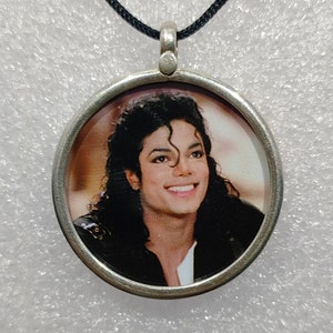 Op de afbeelding: Een zilveren ronde hangerketting met een zwart koord. De hanger heeft een kleurenfoto van Michael Jackson.