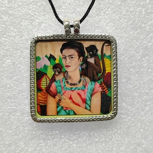 Puede incluir: Un collar con un colgante enmarcado en plata que presenta un retrato colorido de Frida Kahlo con dos monos. El retrato está sobre un fondo verde y amarillo.