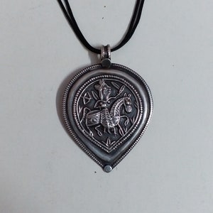 May include: A silver pendant necklace with a black cord. The pendant is shaped like a teardrop and features an intricate design of a person riding a horse.