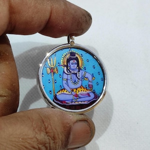 May include: A silver pendant with a blue background and a colorful image of the Hindu deity Shiva. The pendant features a silver border and a loop for attaching a chain.