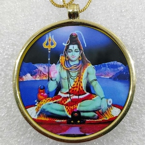 Peut inclure: Pendentif doré avec une image détaillée d'une divinité. L'image représente une figure assise à la peau bleue, tenant un trident. L'arrière-plan montre un ciel bleu, des montagnes et de l'eau. Le pendentif est rond et suspendu à une chaîne dorée.