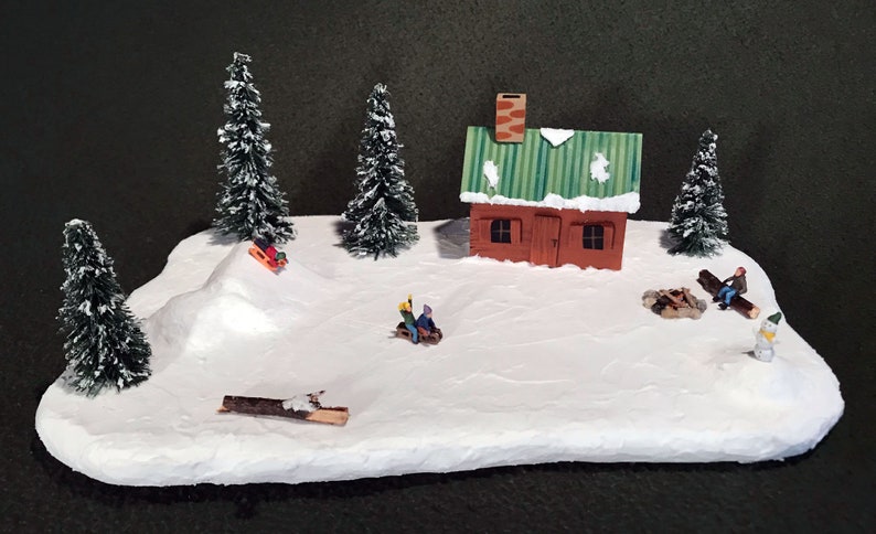 May include: A miniature winter scene with a small red cabin, snow-covered trees, and figures sledding and sitting by a campfire.