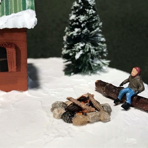 May include: A miniature winter scene with a small red cabin, a snowy evergreen tree, a miniature person sitting on a log by a small campfire, and a snowy white base.