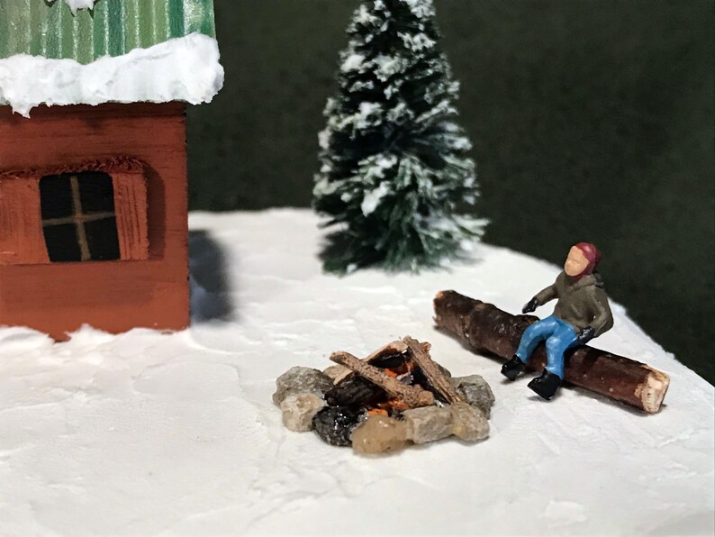 May include: A miniature winter scene with a small red cabin, a snowy evergreen tree, a miniature person sitting on a log by a small campfire, and a snowy white base.