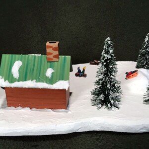 May include: A miniature winter scene with a small brown cabin, green roof, and white snow. There are four small evergreen trees and three figures on sleds.