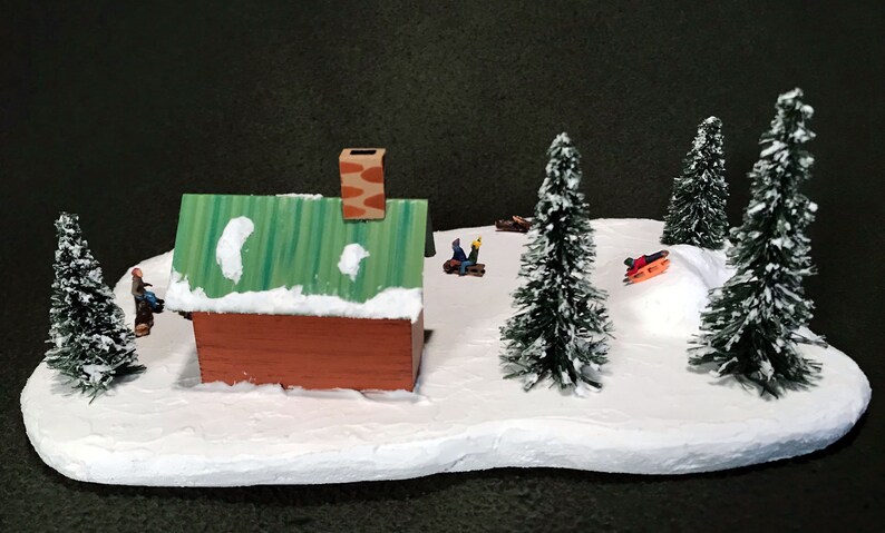 May include: A miniature winter scene with a small brown cabin, green roof, and white snow. There are four small evergreen trees and three figures on sleds.