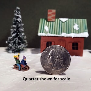 May include: A miniature winter scene with a small red house, two snow-covered pine trees, and two figures on a sled. A US quarter is shown for scale.