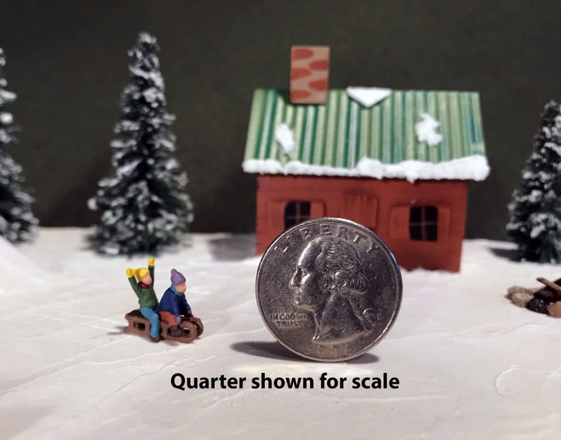 May include: A miniature winter scene with a small red house, two snow-covered pine trees, and two figures on a sled. A US quarter is shown for scale.