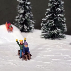 May include: Two miniature figures of children wearing winter clothing are riding a sled down a snowy hill. The scene is set in a winter wonderland with snow-covered trees and a small red cabin in the background.
