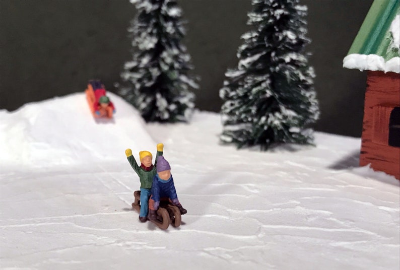 May include: Two miniature figures of children wearing winter clothing are riding a sled down a snowy hill. The scene is set in a winter wonderland with snow-covered trees and a small red cabin in the background.