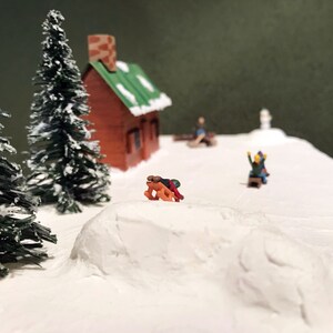 May include: A miniature winter scene with a small brown cabin, two evergreen trees, and figures sledding down a snowy hill.