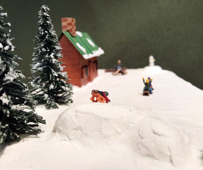 May include: A miniature winter scene with a small brown cabin, two evergreen trees, and figures sledding down a snowy hill.