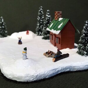 May include: A miniature winter scene with a small red cabin, snow-covered trees, and figures of people playing in the snow. A snowman stands in the foreground.