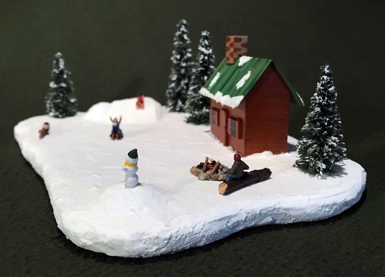 May include: A miniature winter scene with a small red cabin, snow-covered trees, and figures of people playing in the snow. A snowman stands in the foreground.
