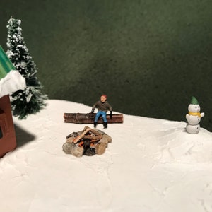 May include: A miniature winter scene with a small wooden cabin, a snowman, a miniature person sitting on a log by a campfire, and a snowy background.