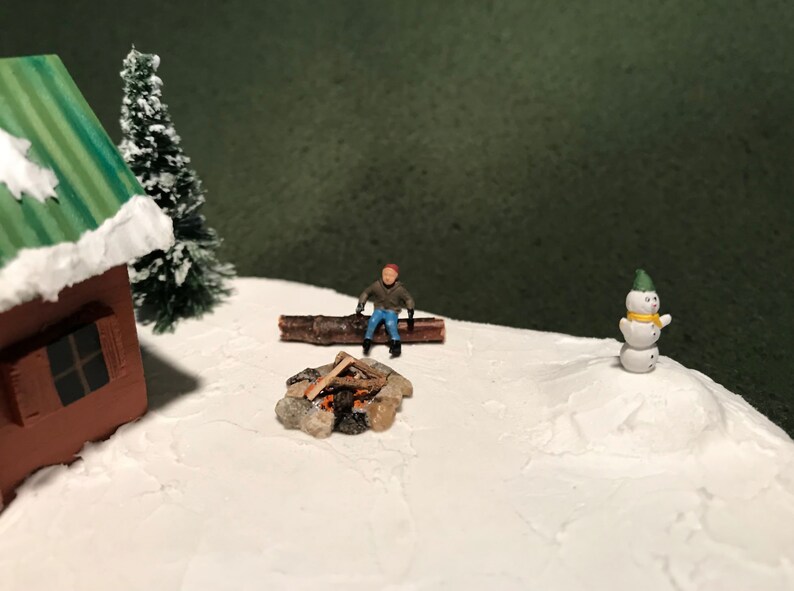 May include: A miniature winter scene with a small wooden cabin, a snowman, a miniature person sitting on a log by a campfire, and a snowy background.