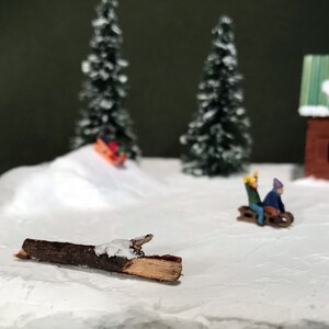 May include: A miniature winter scene with a small wooden cabin, two evergreen trees, and a snow-covered hill. Two figures are riding a sled down the hill, and a small log lies on the snow.