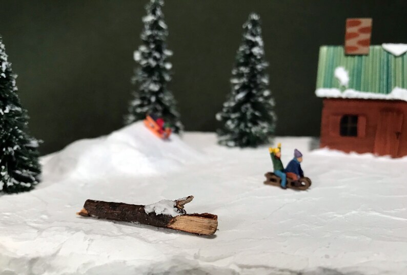 May include: A miniature winter scene with a small wooden cabin, two evergreen trees, and a snow-covered hill. Two figures are riding a sled down the hill, and a small log lies on the snow.