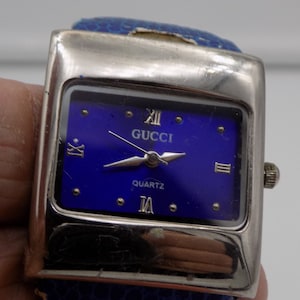 Può includere: Un orologio da polso Gucci al quarzo argentato e blu con quadrante quadrato e cinturino in pelle blu.