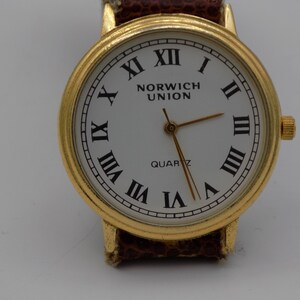 Può includere: Un orologio da polso color oro con quadrante bianco e indici orari in numeri romani neri. Il quadrante dell'orologio presenta anche le parole "Norwich Union" e "Quartz".