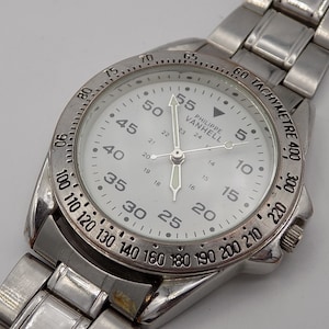 Può includere: Un orologio da polso argentato con quadrante bianco e marcature nere. L'orologio ha una scala tachimetrica attorno al bordo esterno del quadrante. Il quadrante dell'orologio include anche il testo "PHILIPPE VANHELL".