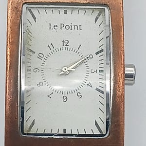 Peut inclure: Une montre rectangulaire avec un boîtier en métal couleur cuivre et un cadran blanc. Le cadran affiche le texte "Le Point" au-dessus des chiffres de 1 à 12. La montre a un bracelet en cuir marron.