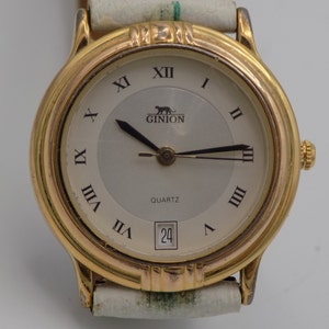 May include: A gold-tone wristwatch with a white face and Roman numeral hour markers. The watch has a date window at the 3 o'clock position and says "GINION QUARTZ" on the face.