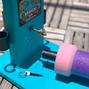May include: A turquoise blue rotisserie with a black handle and a pink and purple foam roller. The rotisserie has a logo that says "The Kraken Mini Rotisserie" with an illustration of an octopus.