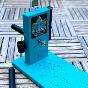 Op de afbeelding: Een turquoise blauwe rotisserie met een zwarte handgreep en een digitale timer. De rotisserie heeft een zwart-wit logo met de tekst "The Kraken Mini Rotisserie".