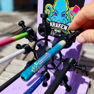 May include: A purple stand with the text "The Kraken Mini Rotisserie" and a blue octopus graphic. The stand holds several colorful, glittery, and patterned rods in place with black adjustable holders.