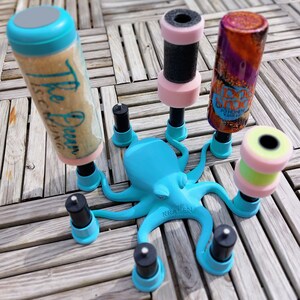 May include: A turquoise octopus-shaped holder with multiple black and pink cylindrical objects. One cylinder has the text "The Ocean is Calling". The holder is on a wooden surface. The text "The Kraken Mini" is on the octopus.